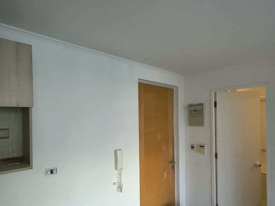 Entrada de un depto con los muros y el cielo recién pintados de blanco, junto a la puerta y el citófono